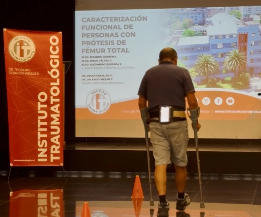 Persona con prótesis de fémur total caminando con bastones y un teléfono fijado al cinturón, durante una evaluación.